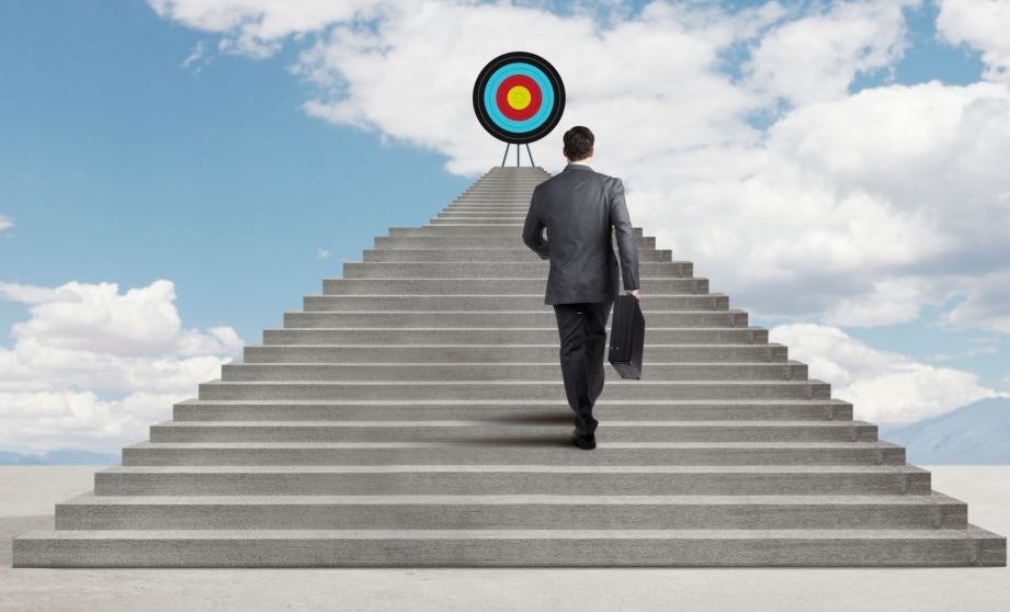 A businessman carries his briefcase while he climbs a set of steps as he strives to reach the top where a target sits. The steps sit under a beautiful, cloud filled sky.