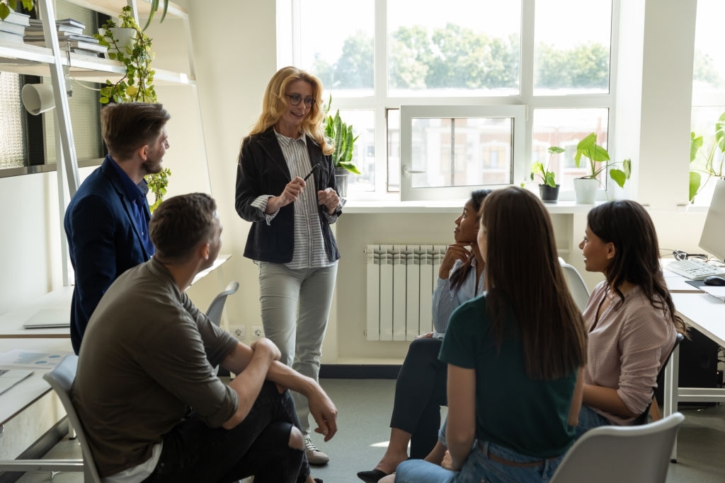 Confident female leader, coach holding briefing for multiracial team employees in office, explaining business strategy, plan to subordinates. Businesswoman at company meeting with diverse colleagues.