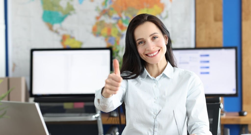Young smiling businesswoman showing thumbs up gesture while sitting at her working table in office. Business consultant and reliable partner concept