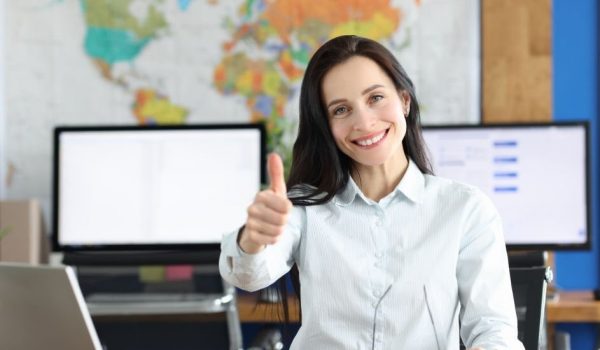 Young smiling businesswoman showing thumbs up gesture while sitting at her working table in office. Business consultant and reliable partner concept