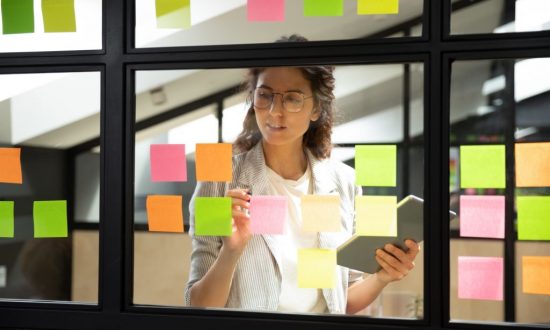 Businesswoman creates priority to-do list standing behind glass wall writes fresh ideas interesting creative thoughts on multicolored post-it sticky notes using tablet having fruitful workday concept