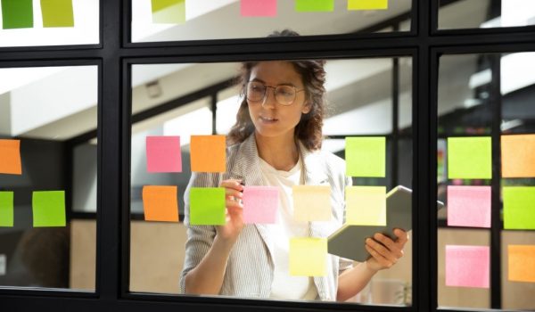 Businesswoman creates priority to-do list standing behind glass wall writes fresh ideas interesting creative thoughts on multicolored post-it sticky notes using tablet having fruitful workday concept