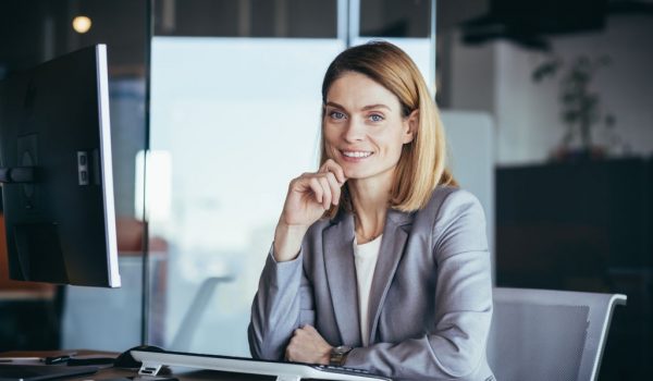 Happy and successful business woman boss working at computer in modern office looking at camera and smiling